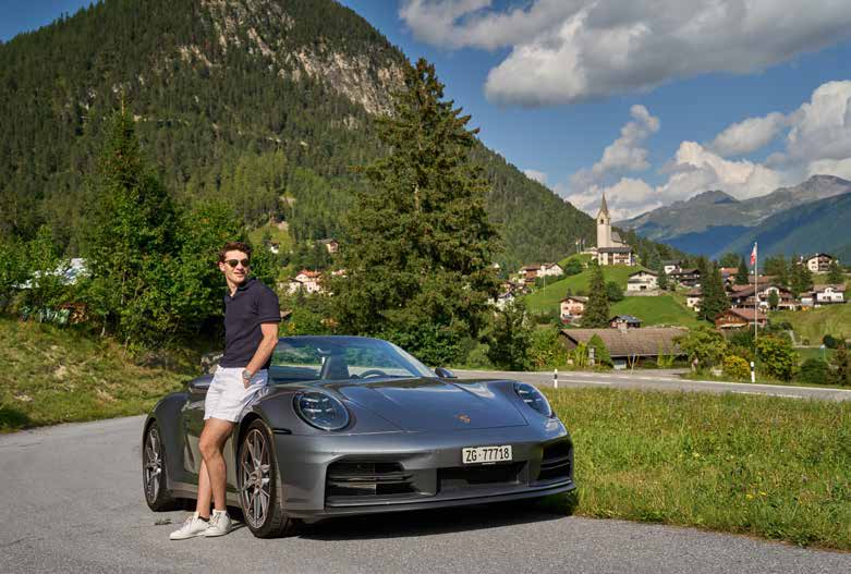 Fahrer lehnt am Porsche 911 Carrera T Cabriolet vor Bergdorf mit Kirche in Graubünden